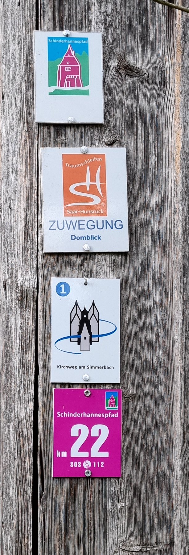 Wegweiserpfosten mit vier Wandermarkierungen: Schinderhannespfad, Traumschleifen Saar-Hunsrück Zuwegung Domblick, Kirchweg am Simmerbach und Kilometerschild Schinderhannespfad km 22